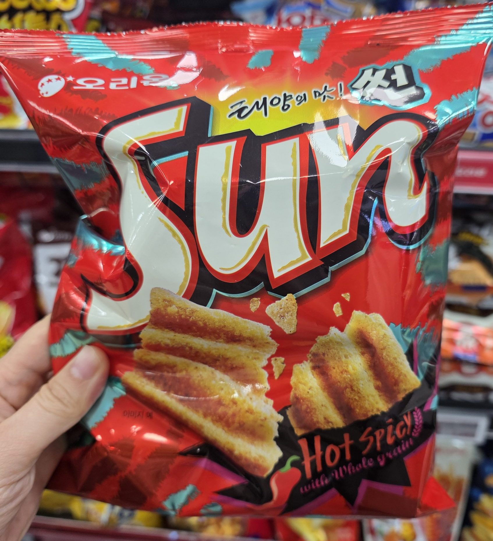 A person holding a red package of Orion Sun Chips in Hot Spicy flavor made with whole grains, displayed in front of a supermarket snack shelf