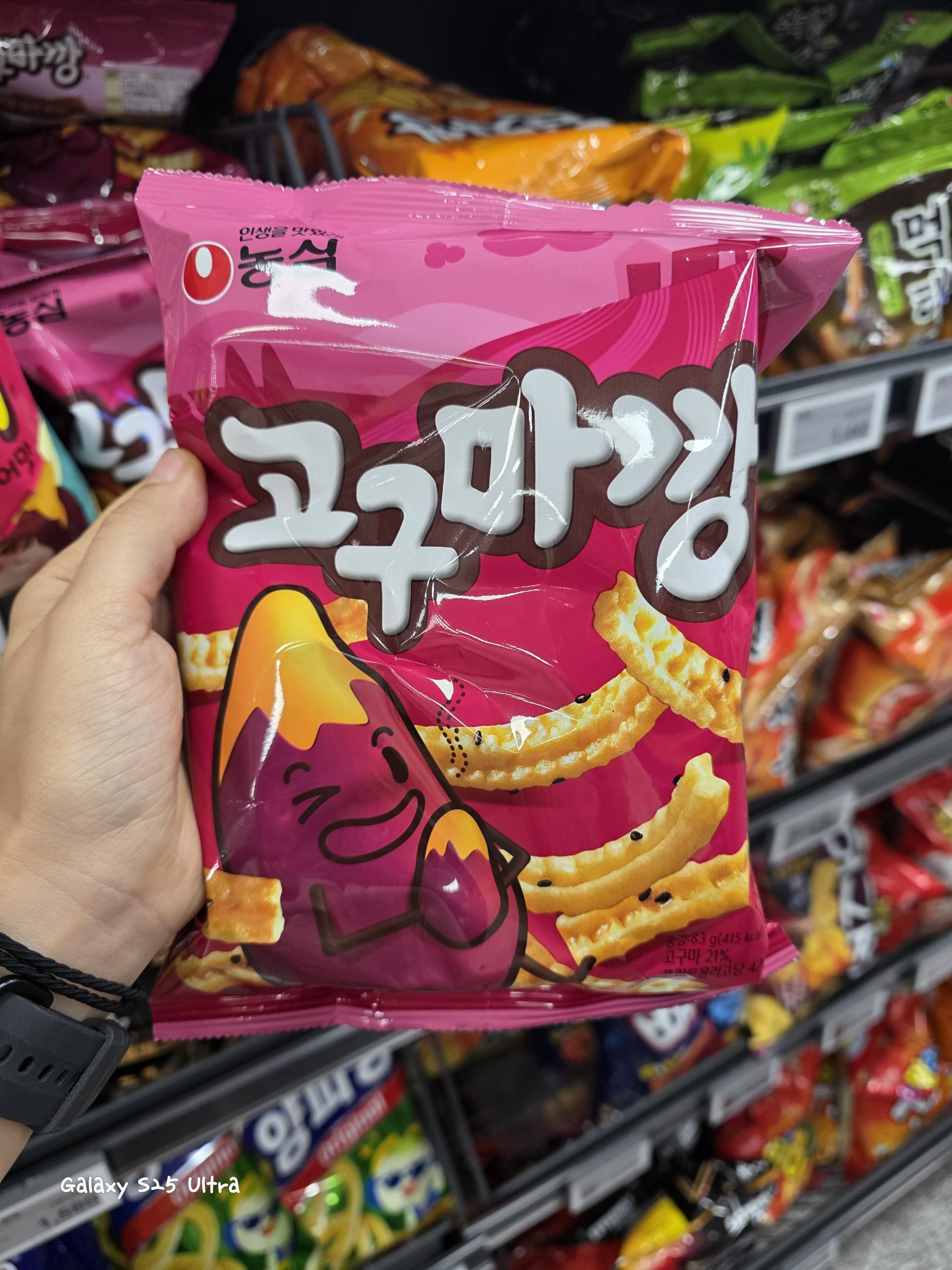 A hand holding a pink bag of Nongshim Goguma Kkang sweet potato flavored snack crackers in a supermarket aisl