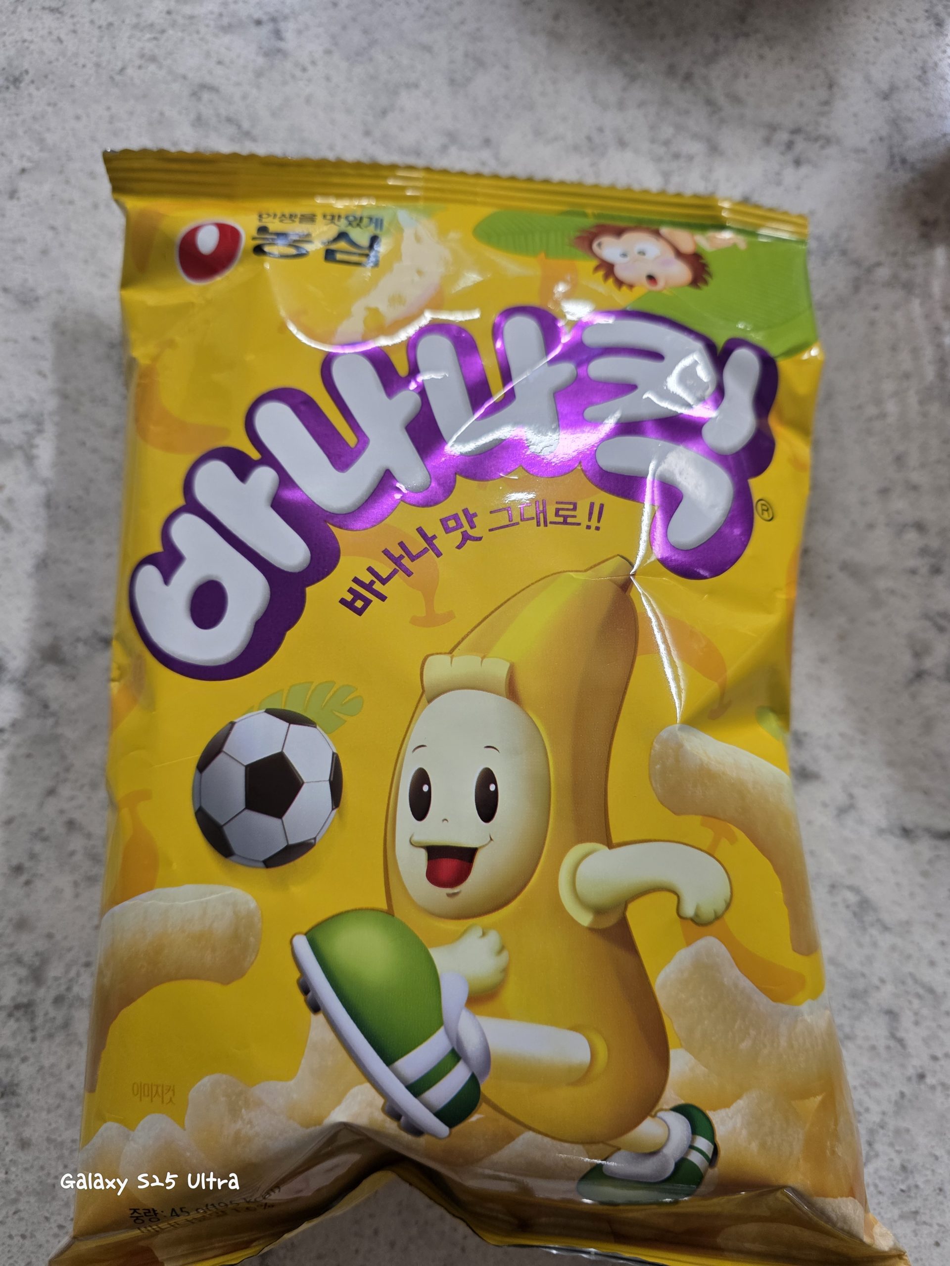 A package of Nongshim Banana Kick (바나나킥), a popular sweet Korean banana-flavored corn puff snack, featuring the playful cartoon banana mascot playing soccer on a bright yellow bag.