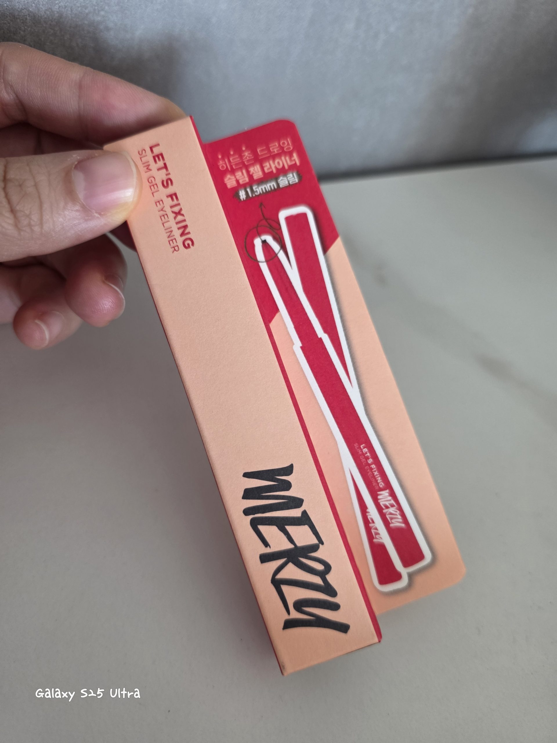 alt="A hand holding the peach-pink and vibrant red packaging of a MERZY eye makeup product. The text 'LET’S FIXING SLIM GEL EYELINER' is printed in red, alongside Korean text '히든존 드로잉 슬림 젤 라이너' and '#1.5mm 슬림'. Features illustrations of the slim gel eyeliner itself with 'MERZY' on the casing, held against a light marble background.