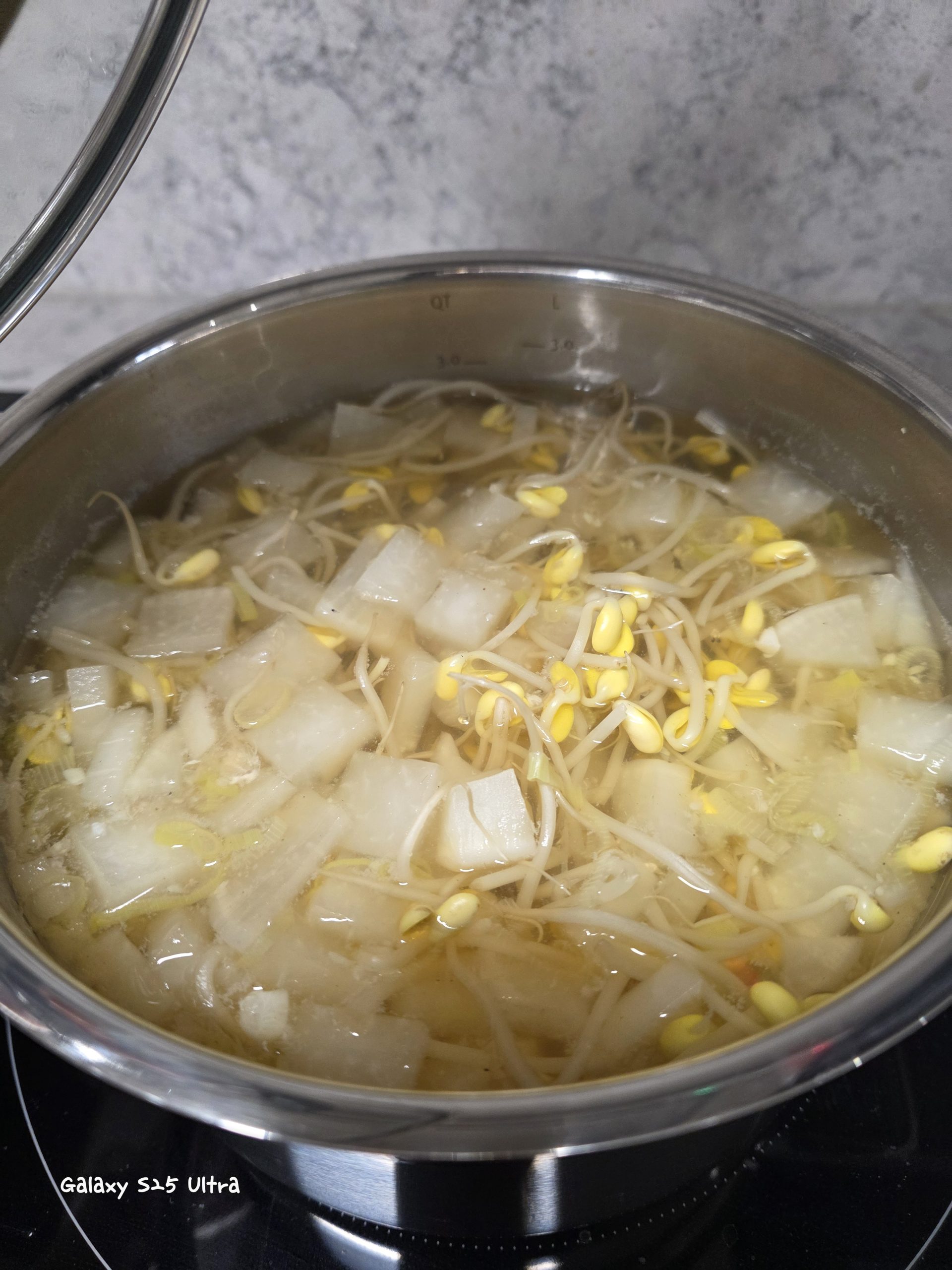A steaming Korean soybean sprout and radish soup (Kongnamul-Muguk) actively boiling in a stainless steel pot on an induction cooktop. The clear broth is densely filled with yellow-headed soybean sprouts and diced white radish cubes, against a blurred kitchen tile background.