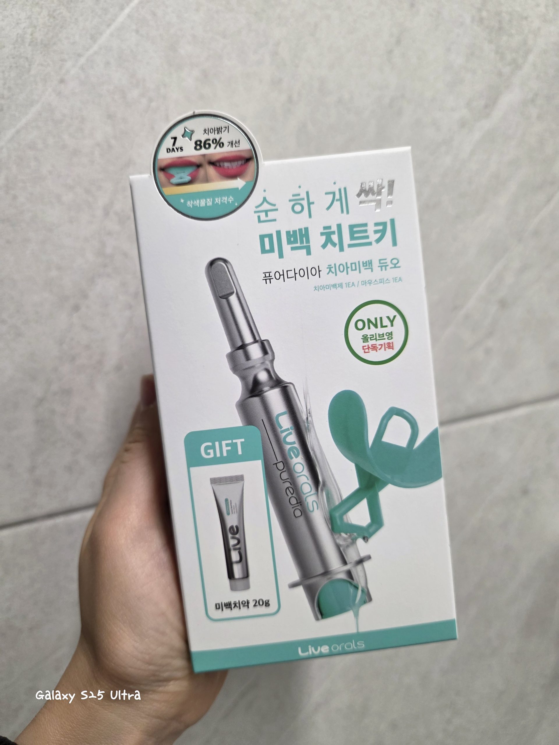 A hand holding the packaging box of Live Orals Pure Dia Teeth Whitening Duo, an Olive Young exclusive set. The white box displays a syringe-style teeth whitening applicator, a green silicone mouth tray, and a bonus 20g whitening toothpaste, with Korean promotional text highlighting 86% brightness improvement in 7 days.