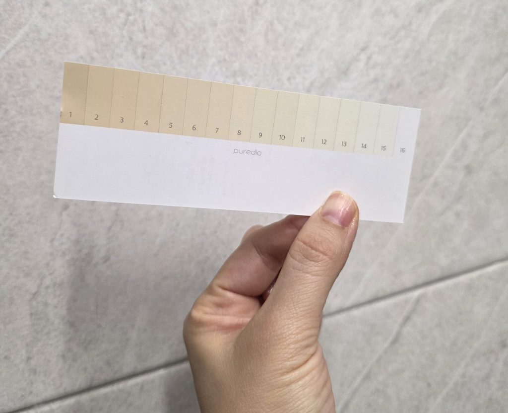 A hand holding a paper teeth shade guide from Pure Dia against a gray tiled wall, featuring a 16-step color gradient scale numbered from dark yellow to bright white to measure whitening progress.
