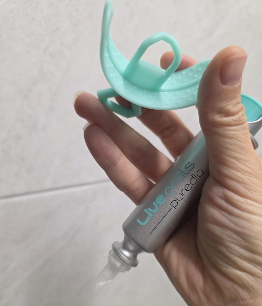 A hand holding the components of the Live Orals Pure Dia teeth whitening kit, featuring a metallic silver whitening gel syringe applicator with a silicone brush tip and a light green silicone mouth tray, shown against a neutral tiled background.