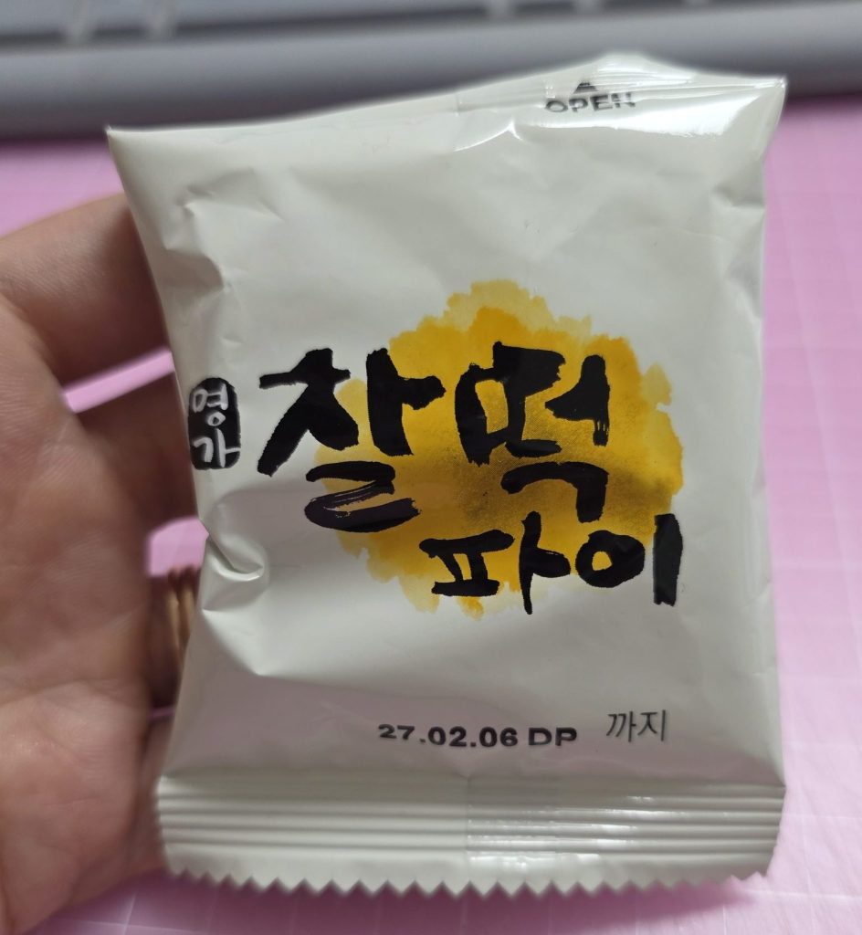 A hand holding an unopened individual package of Lotte Myungga Chal-tteok Pie, featuring bold Korean calligraphy lettering '찰떡파이' over a yellow brushstroke design, with an expiration date printed at the bottom on a pink desk mat.
