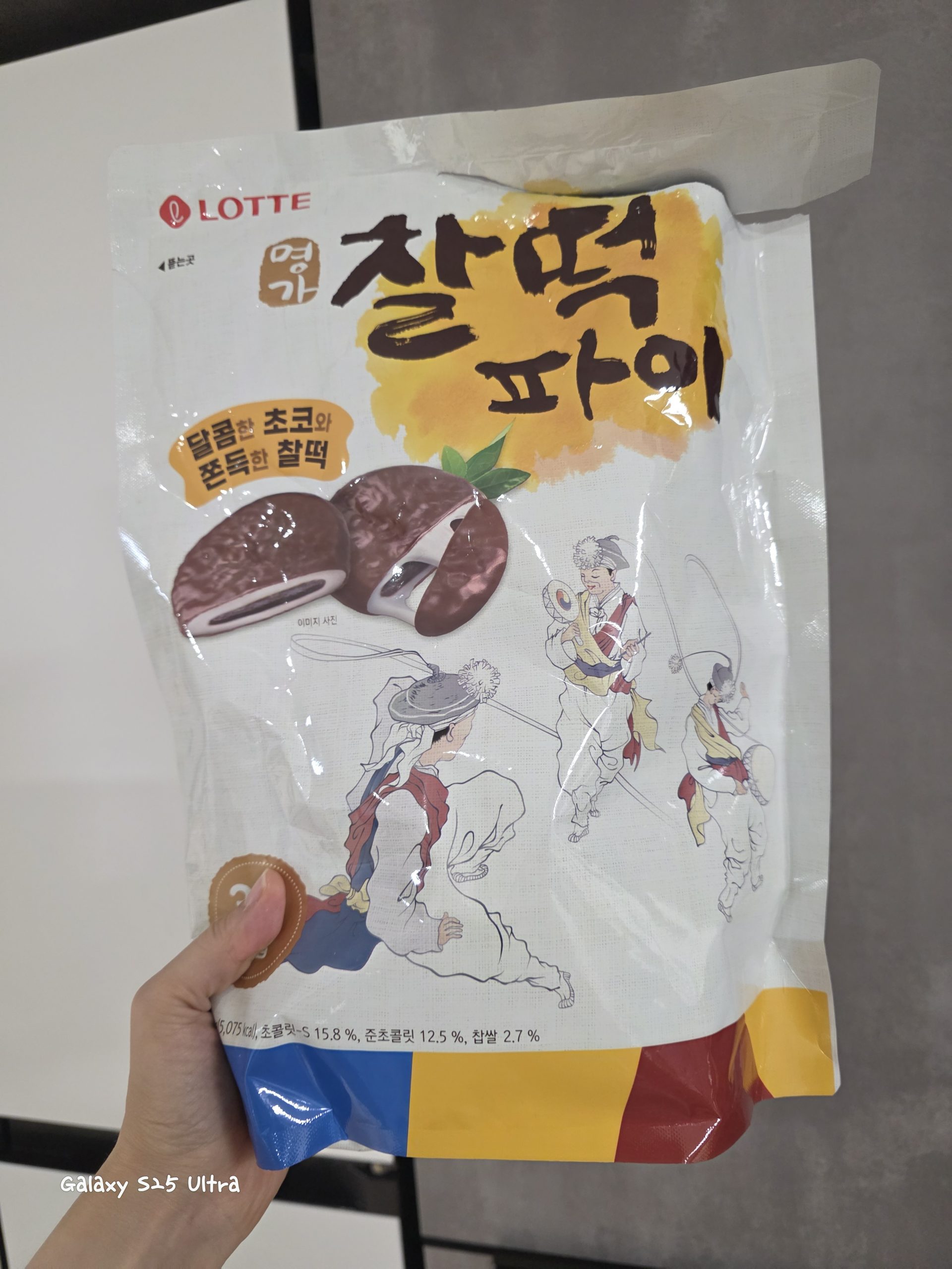 A hand holding a large white bag of Lotte Myungga Chal-tteok Pie (chocolate covered chewy rice cake). The package prominently displays bold calligraphy text '찰떡파이', photos of chocolate-coated glutinous rice pies filled with sweet chocolate cream, and traditional Korean folk art illustrations of Nongak (farmers' music) dancers.
