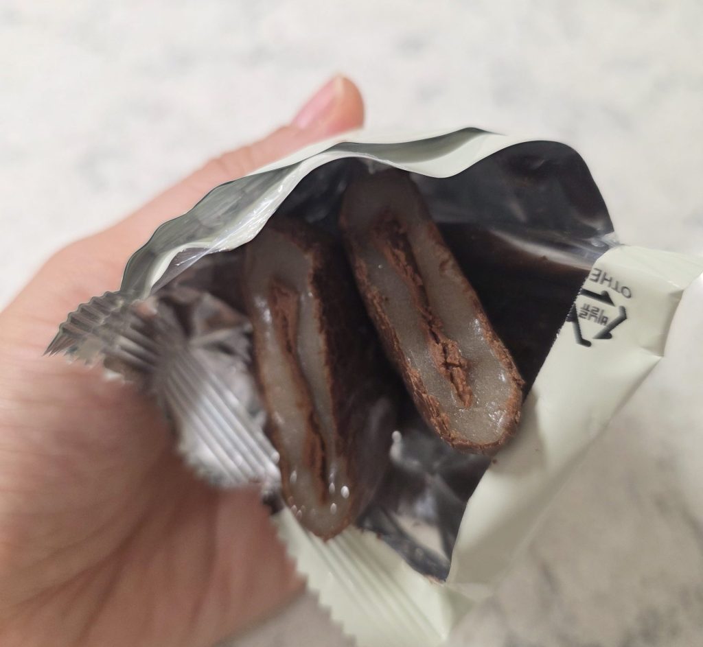 A close-up view of an opened individual wrapper held in hand, revealing a Korean Chal-tteok Pie split in half to show the chewy glutinous rice cake layer coated in chocolate and filled with rich chocolate cream.