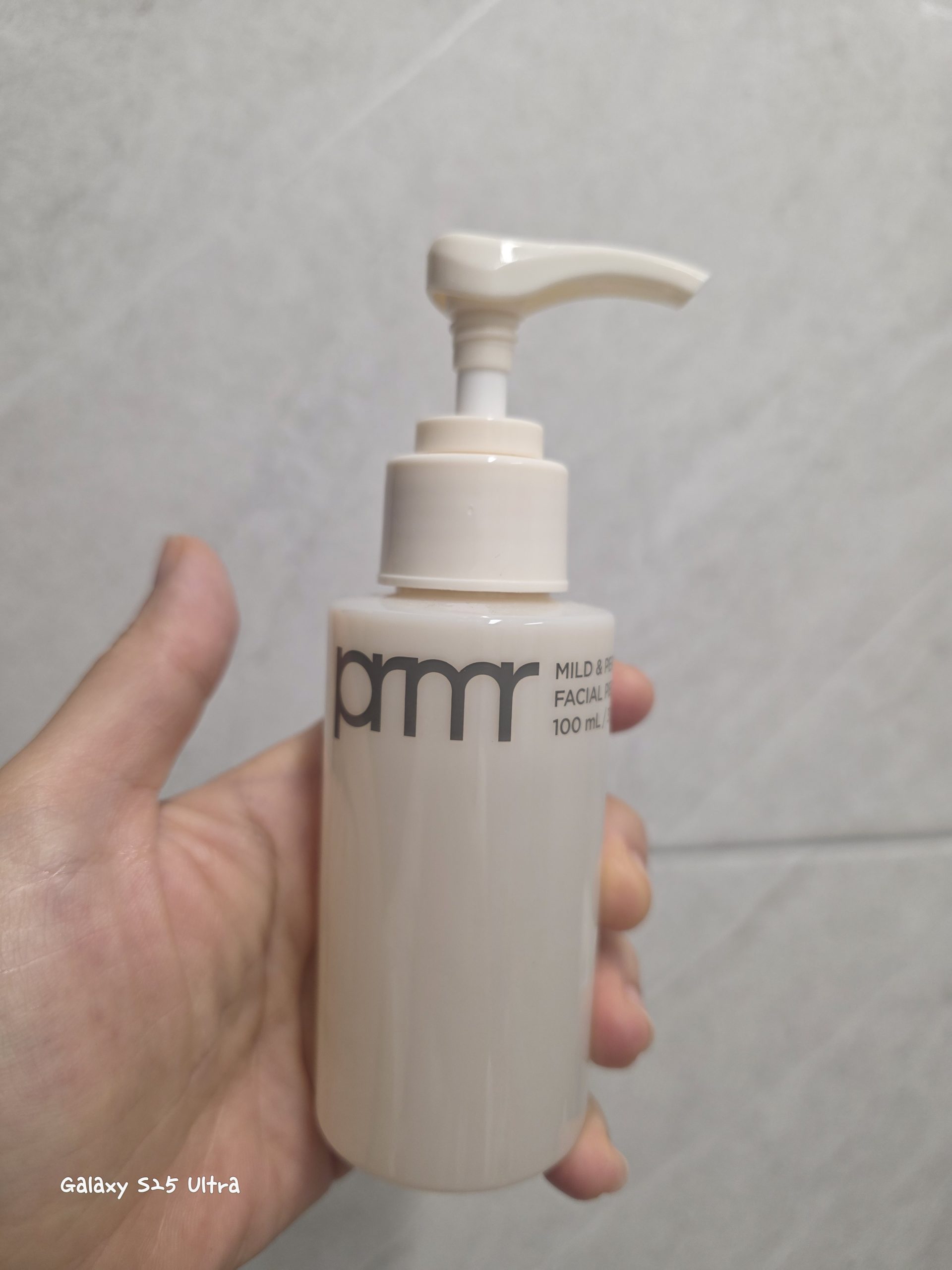 A hand holding a 100ml pump bottle of pmr Mild Facial cleanser against a light gray tiled wall, featuring a clean minimalist beige-and-white design.