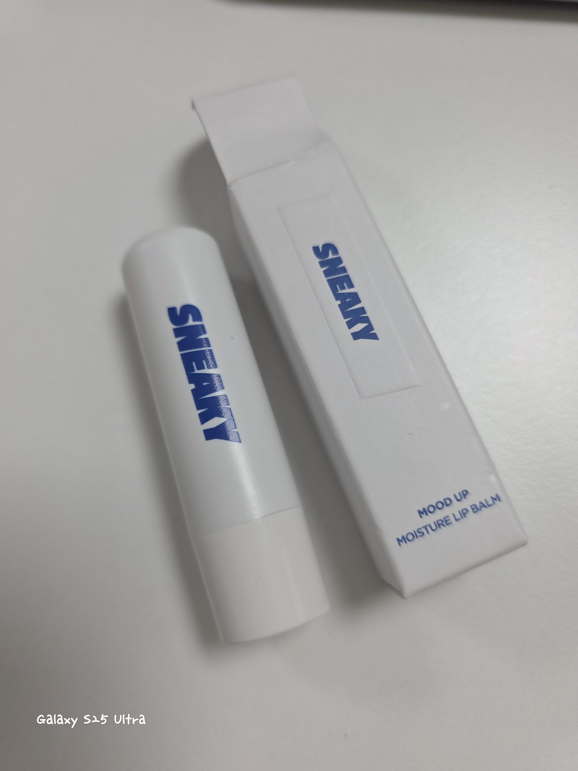 Sneaky Mood Up Moisture Lip Balm stick and its white paper packaging box with bold blue branding, displayed on a clean white desk surface.
