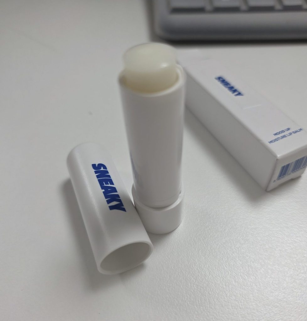 An unboxed Sneaky Mood Up Moisture Lip Balm opened to reveal its translucent moisturizing balm stick, with the cap resting beside it and the packaging box in the soft-focus background on a desk.