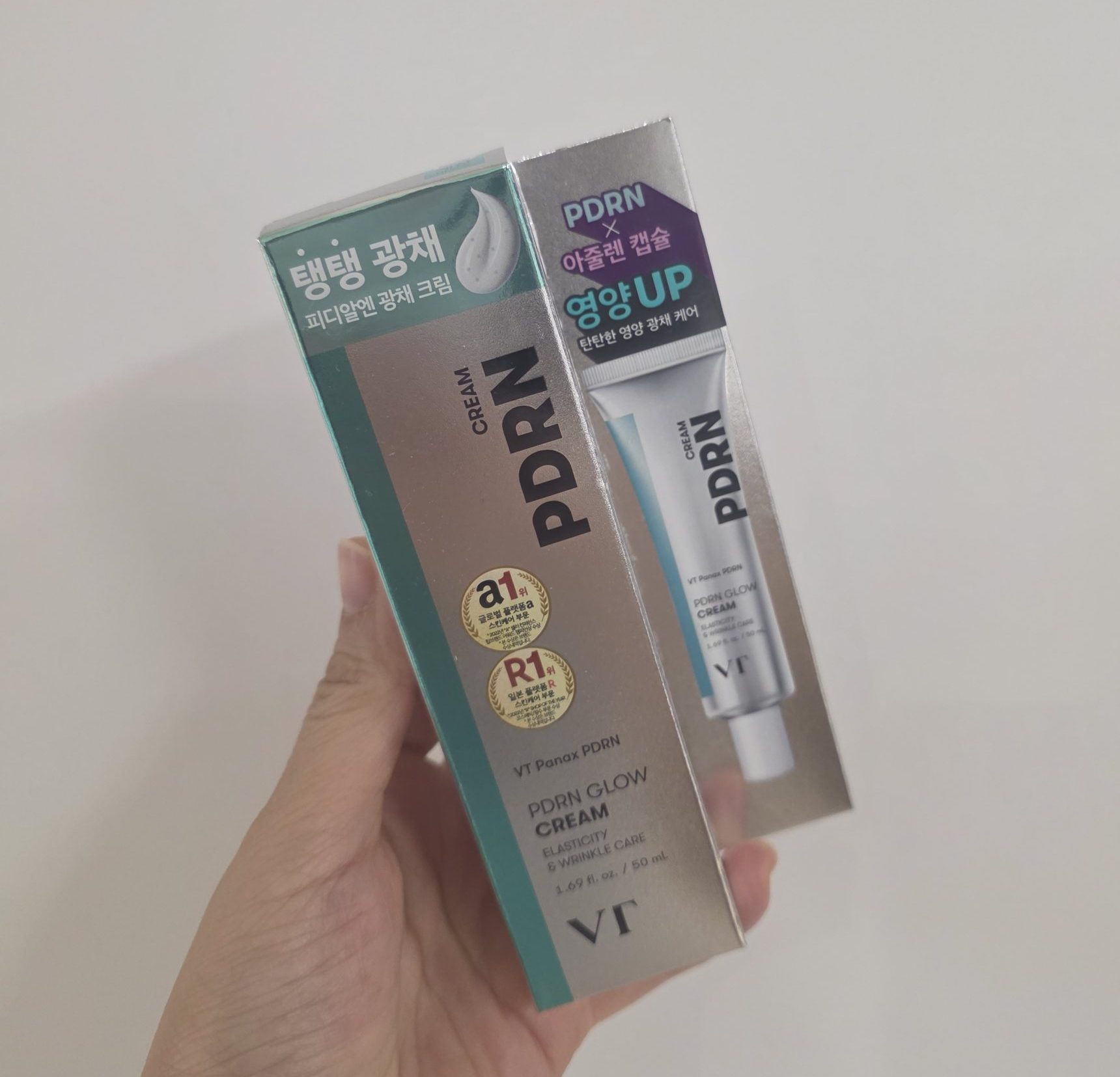 Hand holding a box of VT PDRN Glow Cream for elasticity and wrinkle care in front of a white wall