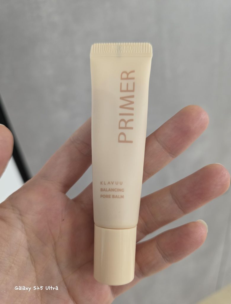 Hand holding a tube of Klavuu Balancing Pore Balm Primer in front of a neutral gray background.