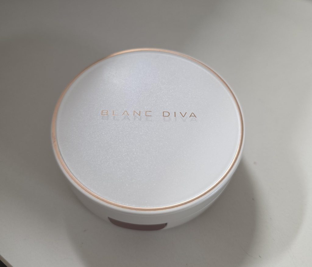 A close-up view of a circular, pearl-white 'BLANC DIVA' cushion compact case resting on a desk. The compact features metallic gold trim and the brand name in gold lettering on a slightly shimmering, textured white lid.
